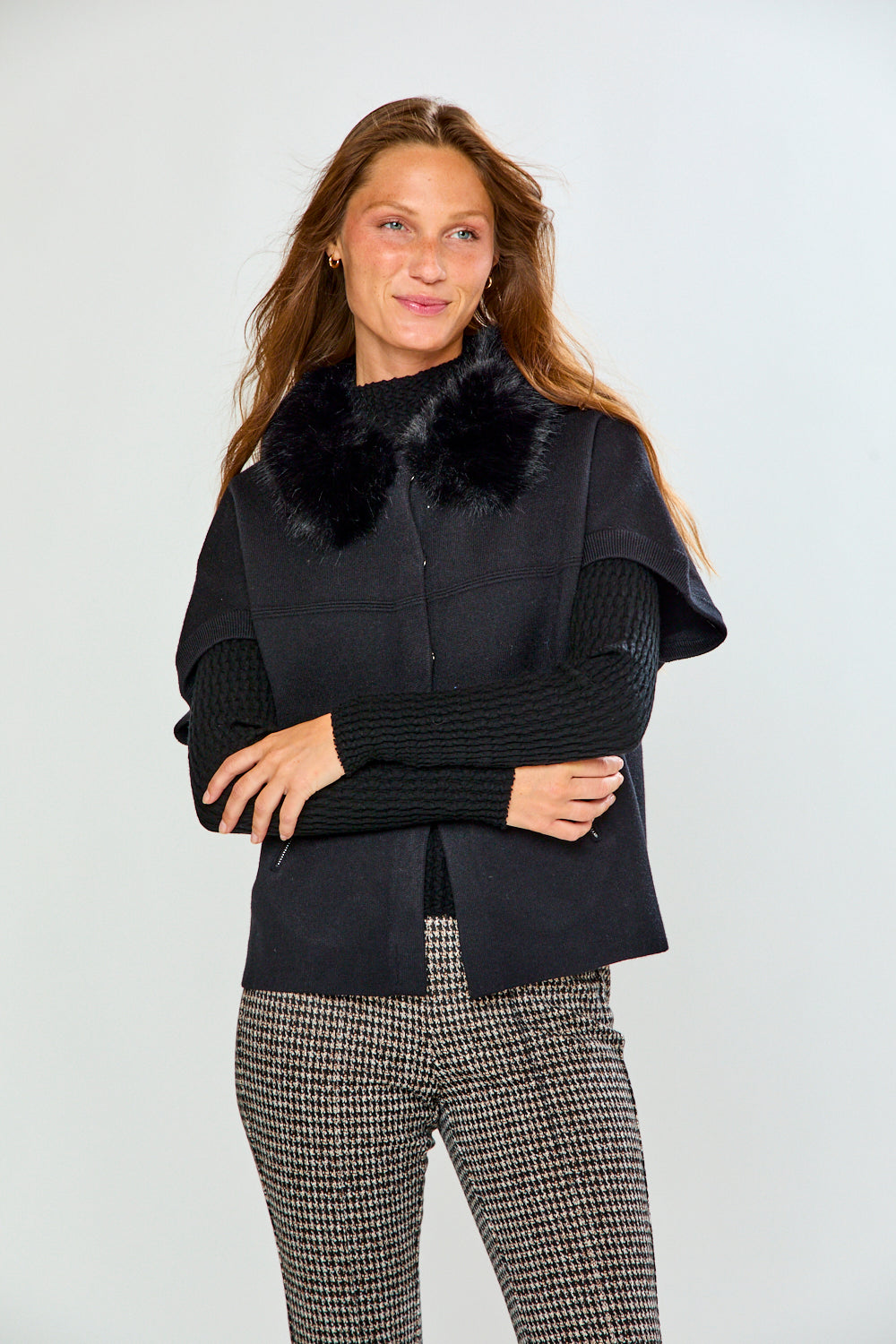 A woman modeling a black sleeveless sweater with a fluffy black faux-fur collar, worn over a black ribbed long-sleeve top and paired with black-and-white houndstooth pants, against a light gray studio background.