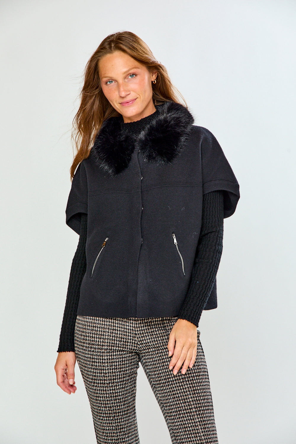 A woman modeling a black sleeveless sweater with a fluffy black faux-fur collar, worn over a black ribbed long-sleeve top and paired with black-and-white houndstooth pants, against a light gray studio background.