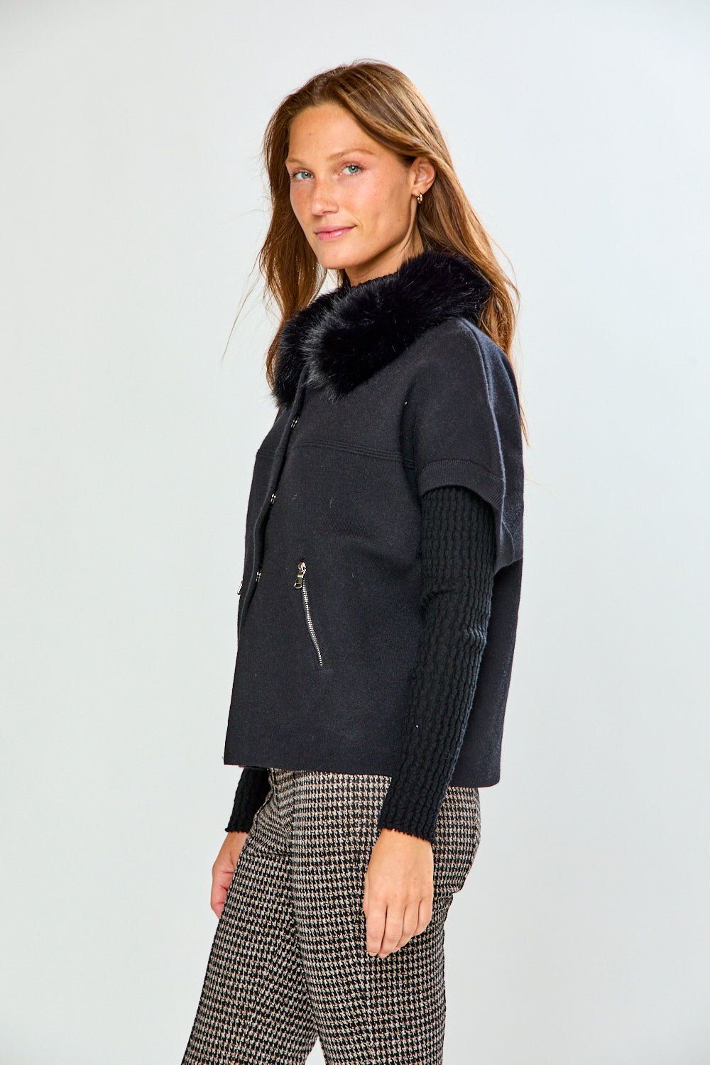 A woman modeling a black sleeveless sweater with a fluffy black faux-fur collar, worn over a black ribbed long-sleeve top and paired with black-and-white houndstooth pants, against a light gray studio background.