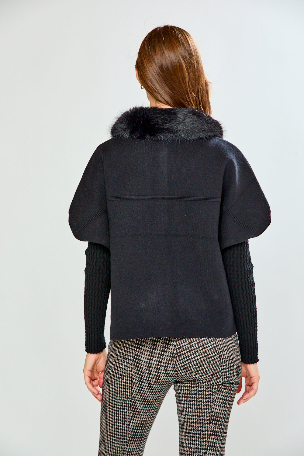 A woman modeling a black sleeveless sweater with a fluffy black faux-fur collar, worn over a black ribbed long-sleeve top and paired with black-and-white houndstooth pants, against a light gray studio background.