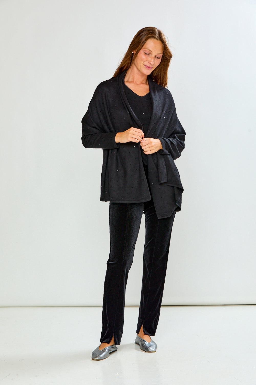 A woman modeling a black shawl with subtle silver rhinestone embellishments over a black top and black velvet pants, photographed against a light gray studio background.