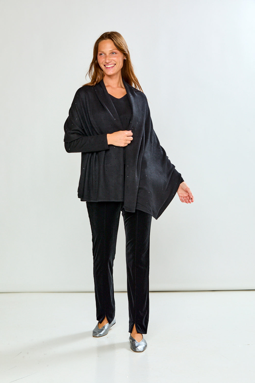 A woman modeling a black shawl with subtle silver rhinestone embellishments over a black top and black velvet pants, photographed against a light gray studio background.