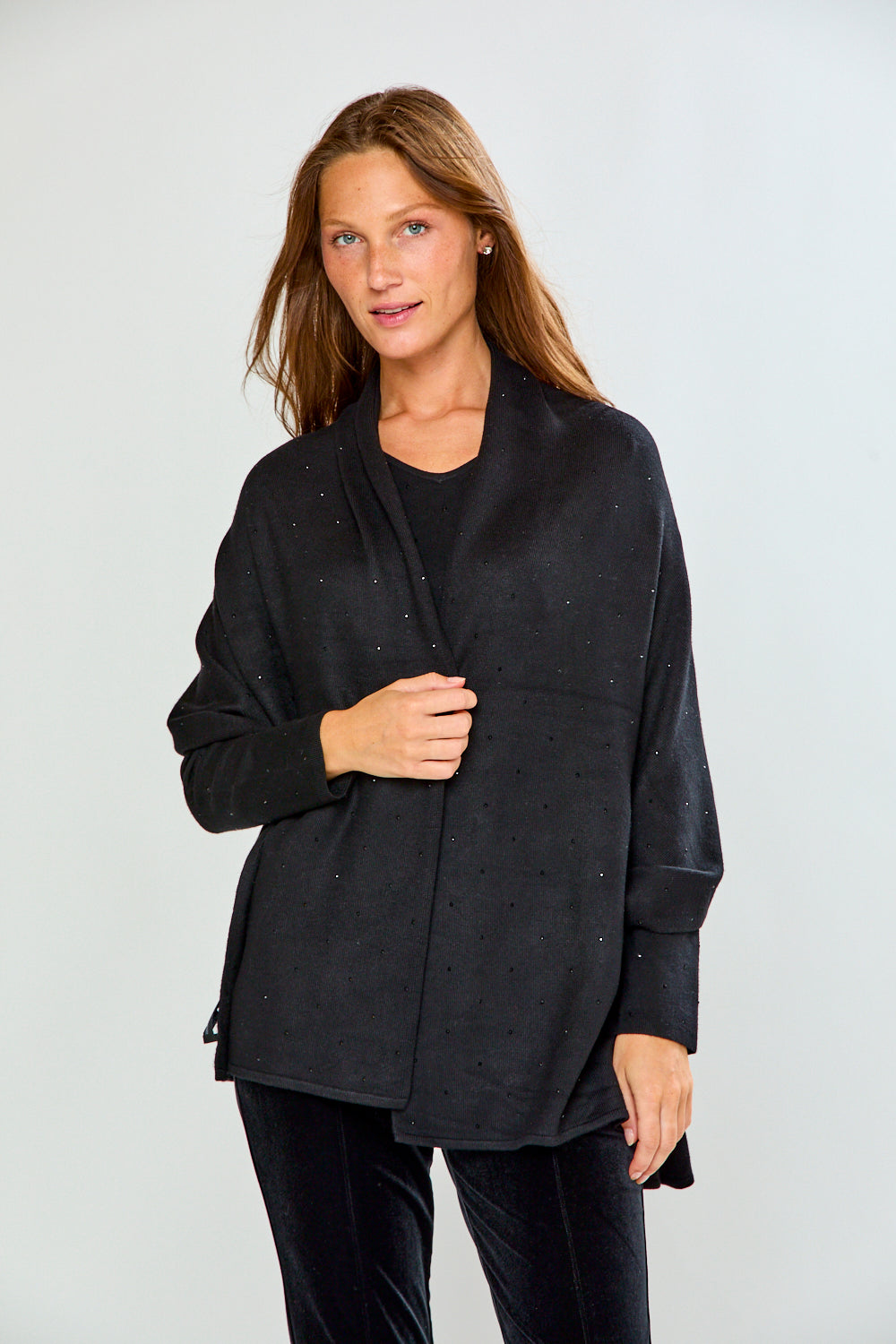 A woman modeling a black shawl with subtle silver rhinestone embellishments over a black top and black velvet pants, photographed against a light gray studio background.