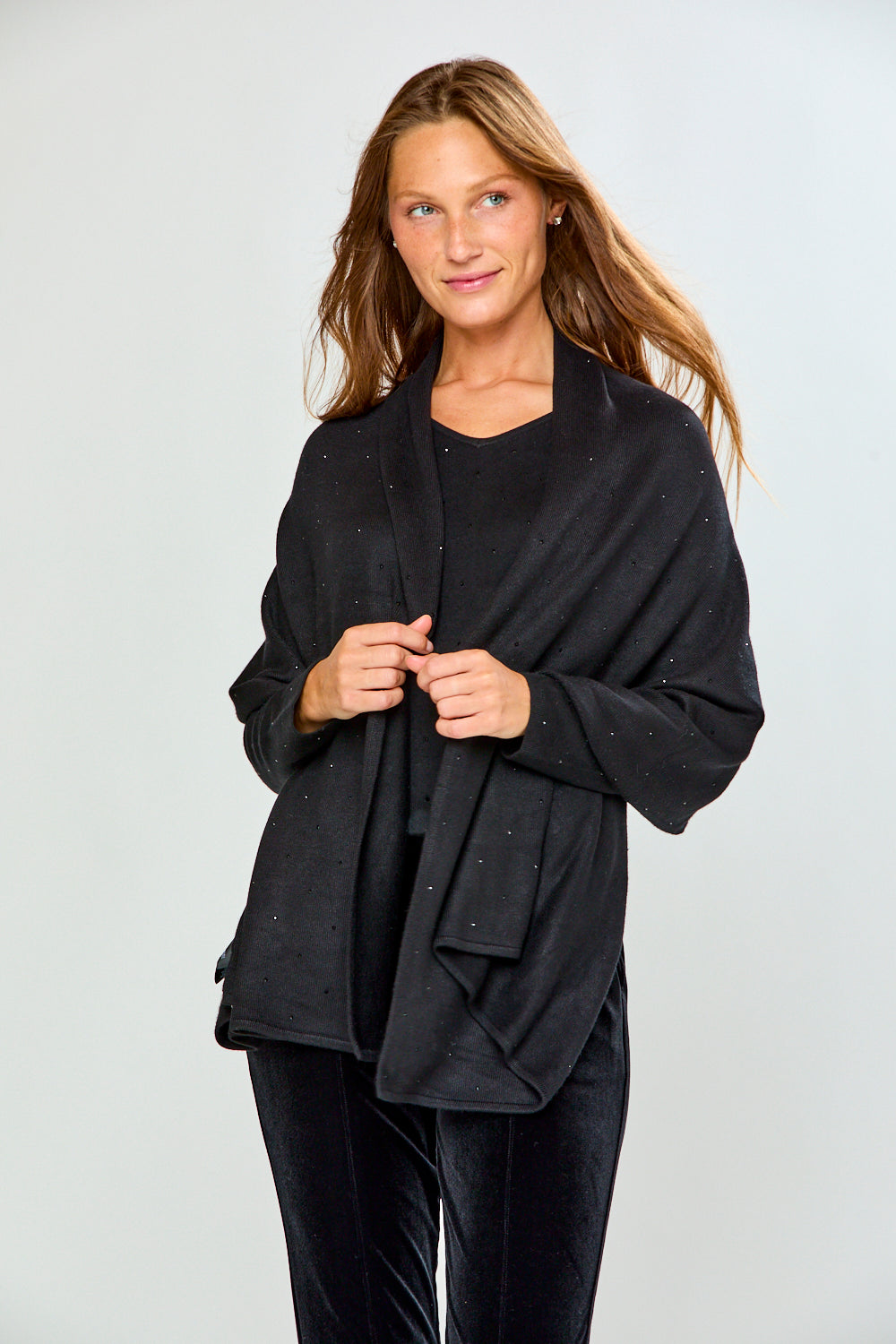 A woman modeling a black shawl with subtle silver rhinestone embellishments over a black top and black velvet pants, photographed against a light gray studio background.