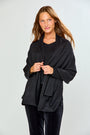 A woman modeling a black shawl with subtle silver rhinestone embellishments over a black top and black velvet pants, photographed against a light gray studio background.