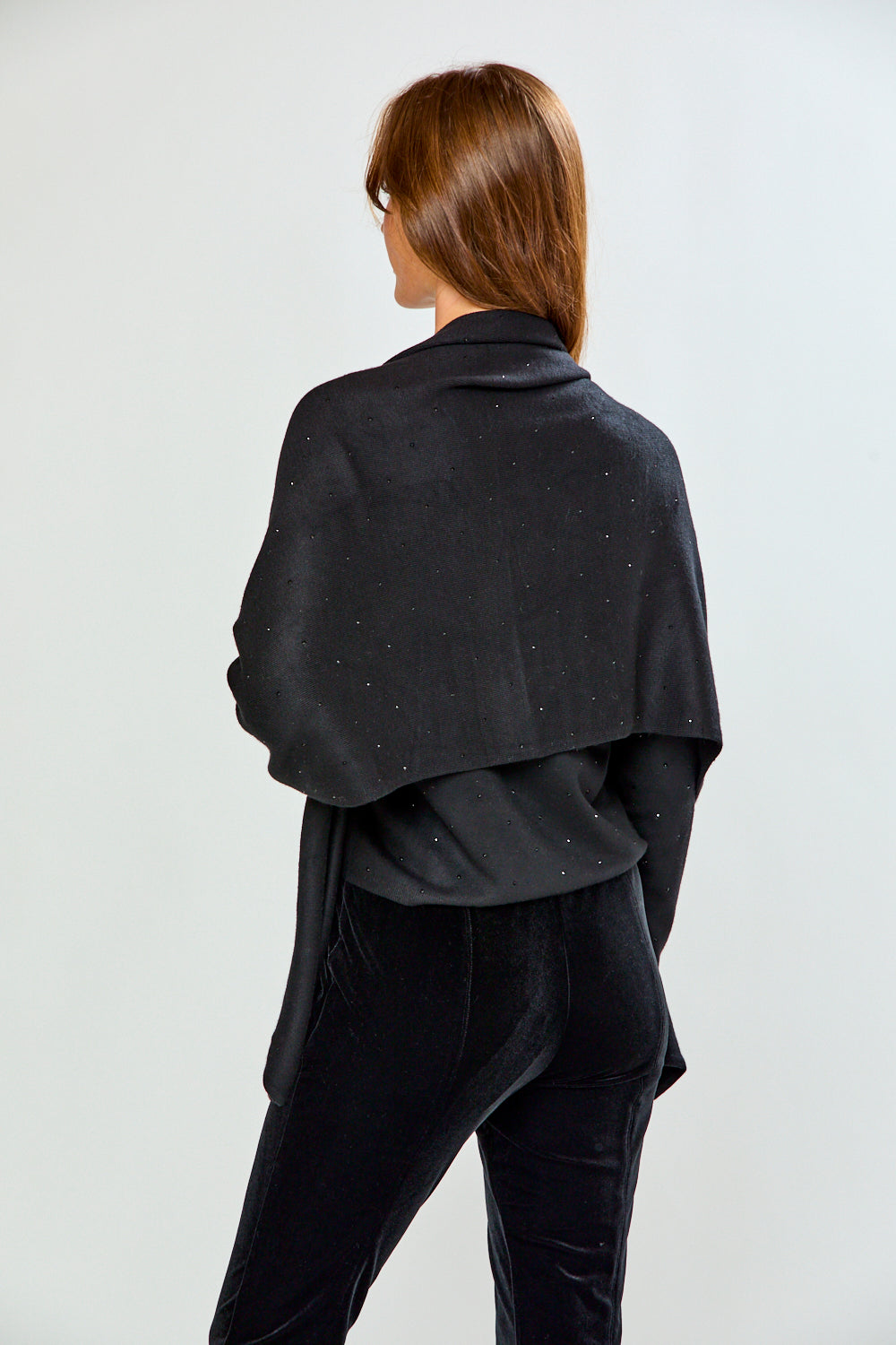 A woman modeling a black shawl with subtle silver rhinestone embellishments over a black top and black velvet pants, photographed against a light gray studio background.