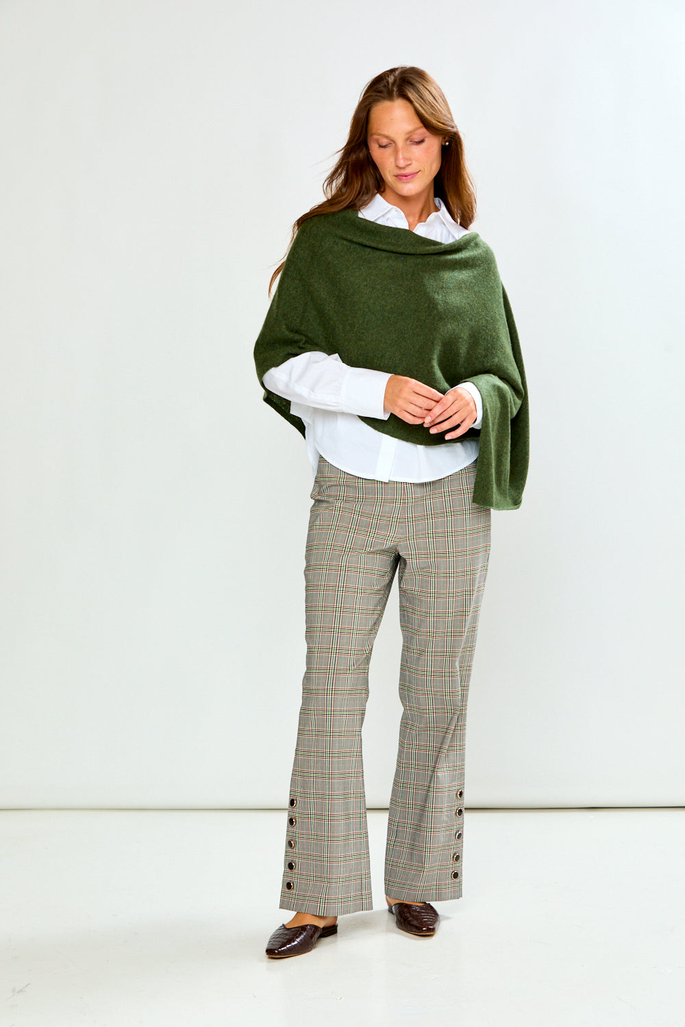 A woman modeling an olive green poncho draped over her shoulders, styled with a white collared shirt and beige plaid trousers, against a plain light gray background.
