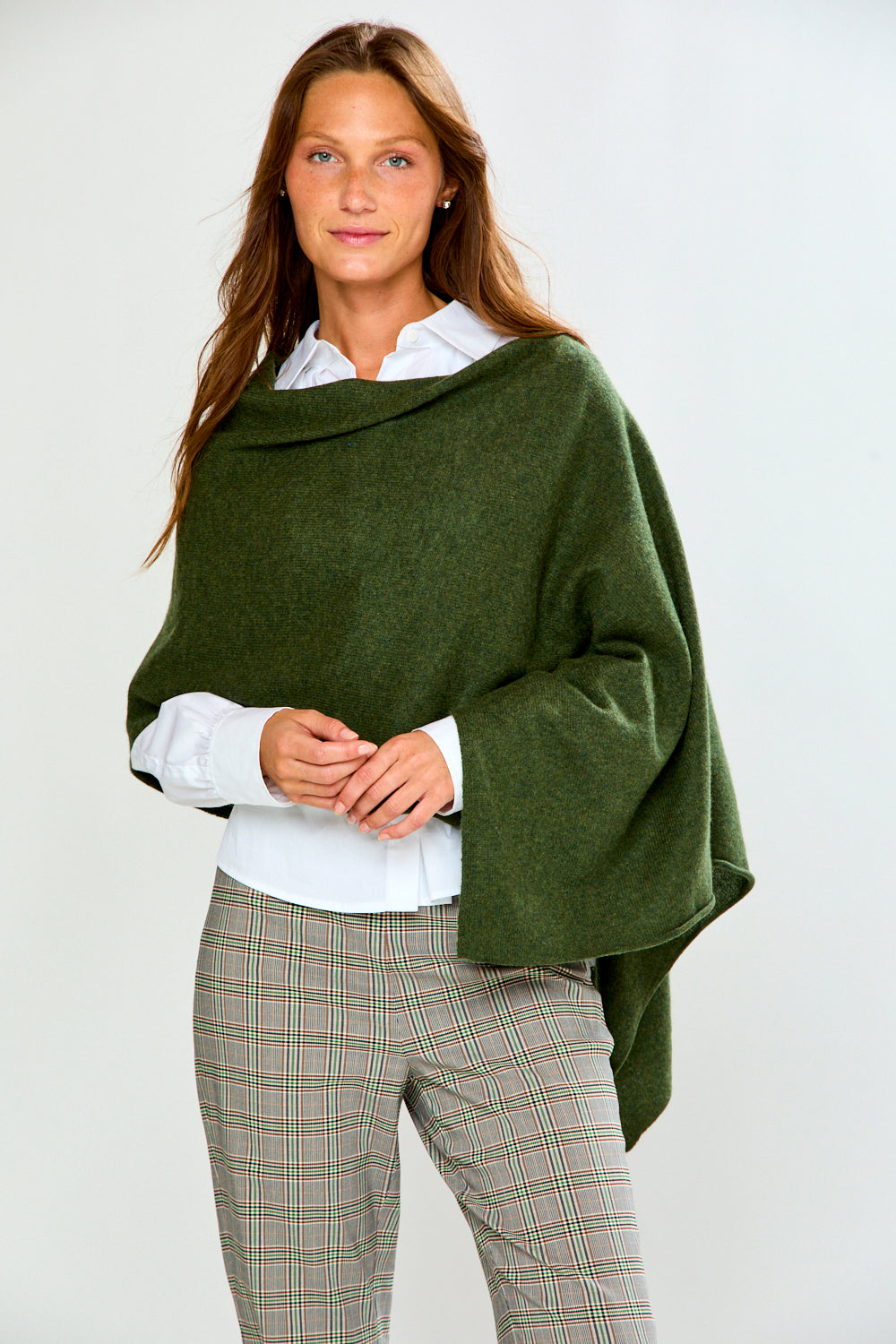 A woman modeling an olive green poncho draped over her shoulders, styled with a white collared shirt and beige plaid trousers, against a plain light gray background.