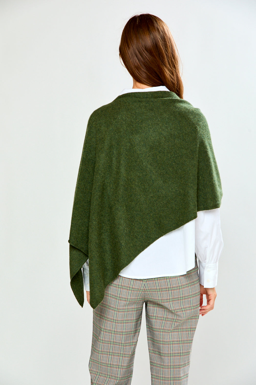 A woman modeling an olive green poncho draped over her shoulders, styled with a white collared shirt and beige plaid trousers, against a plain light gray background.