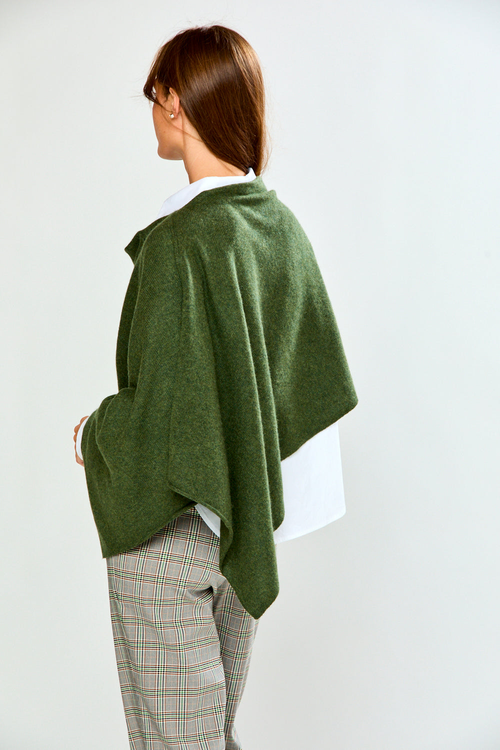 A woman modeling an olive green poncho draped over her shoulders, styled with a white collared shirt and beige plaid trousers, against a plain light gray background.