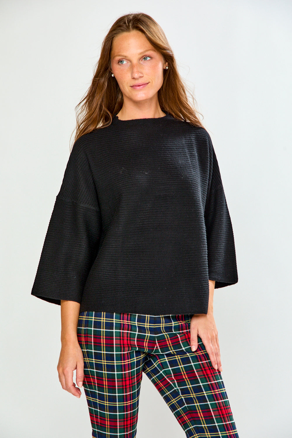 A woman modeling a black textured sweater with wide three-quarter sleeves and red, green, and yellow plaid pants against a light gray studio background.