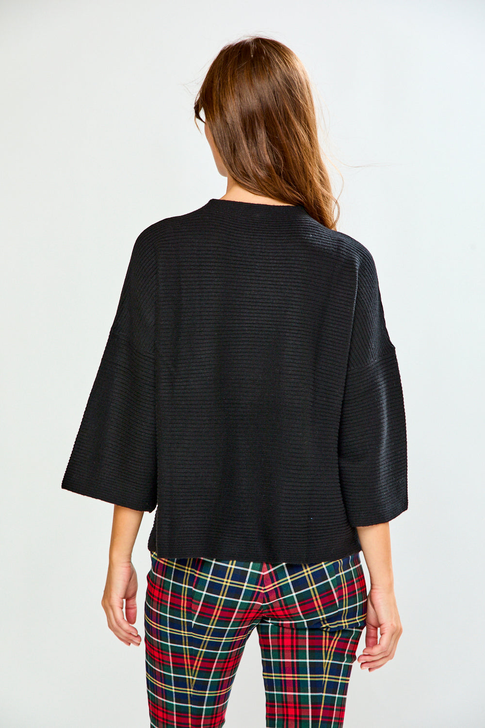 A woman modeling a black textured sweater with wide three-quarter sleeves and red, green, and yellow plaid pants against a light gray studio background.