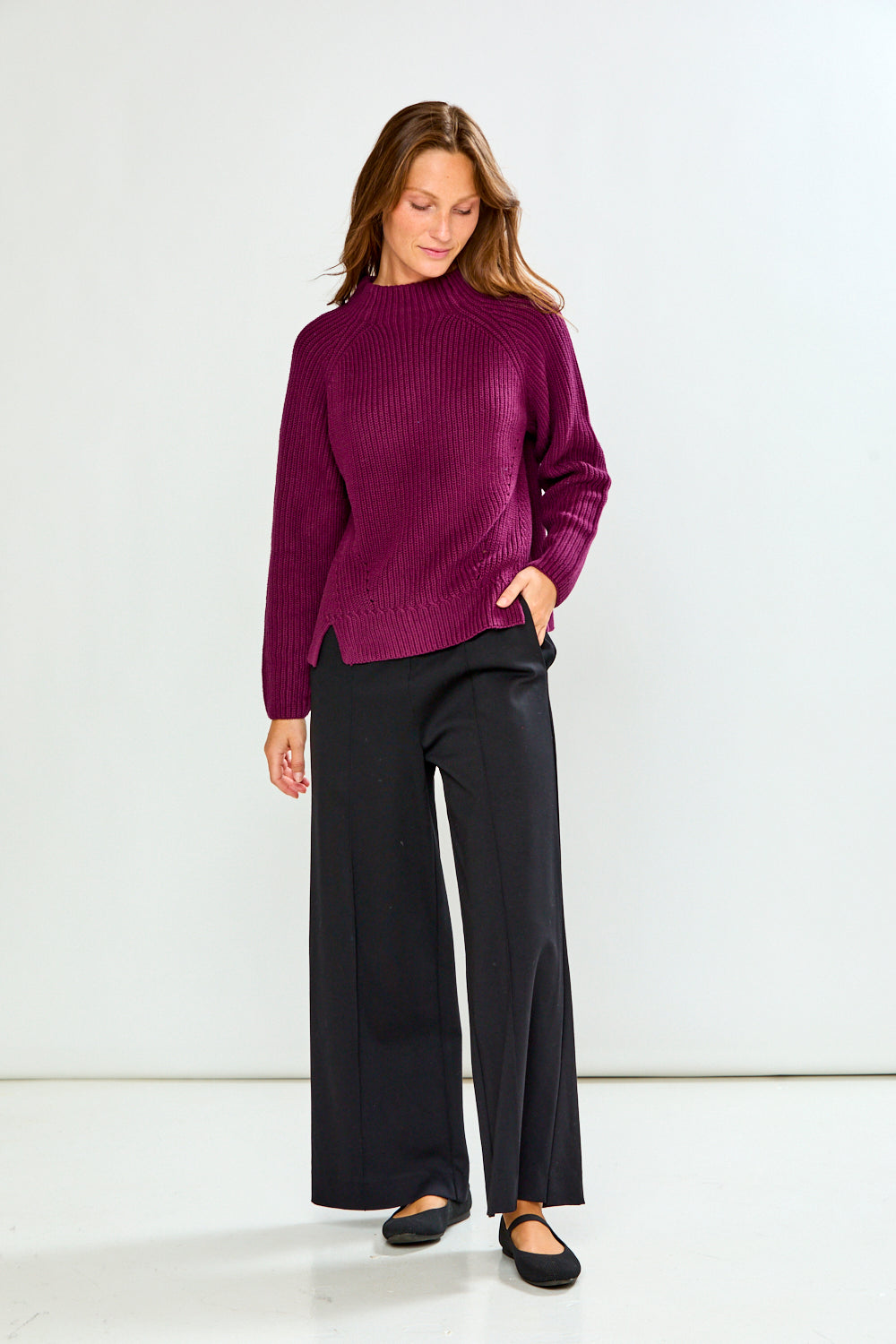 A woman modeling a ribbed knit sweater in deep purple with wide-leg black pants and black flats, standing against a plain white studio background.