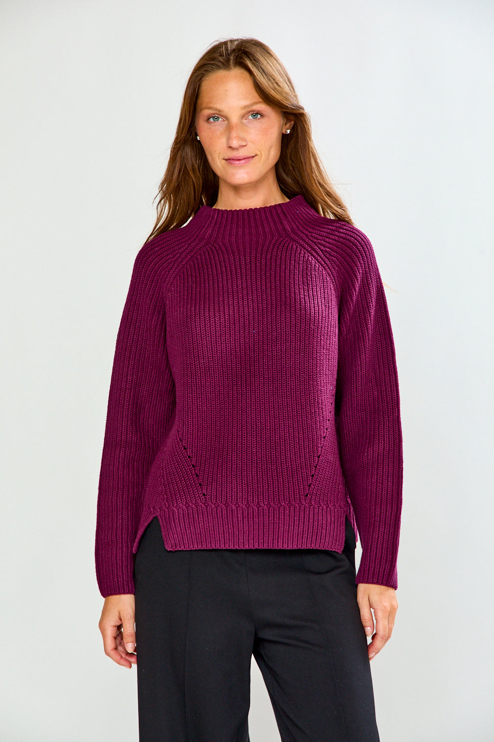 A woman modeling a ribbed knit sweater in deep magenta-purple with black trousers, standing against a light gray studio background.
