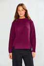 A woman modeling a ribbed knit sweater in deep magenta-purple with black trousers, standing against a light gray studio background.