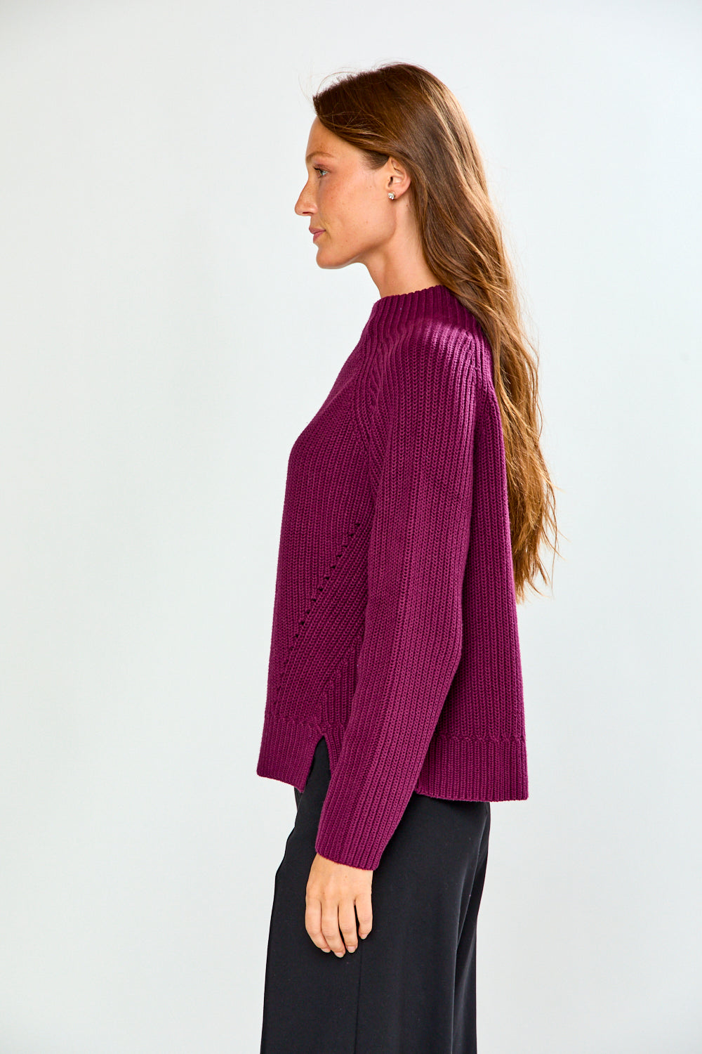 A woman in profile wearing a ribbed knit long-sleeve sweater in deep magenta-purple and black trousers, standing against a light gray studio background.