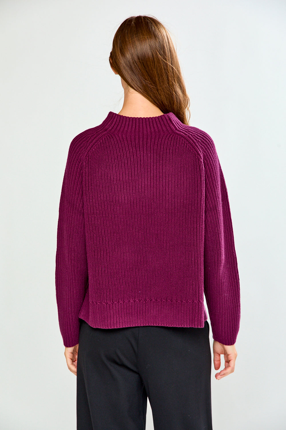 A woman viewed from the back wearing a ribbed knit long-sleeve sweater in deep magenta-purple and black pants, against a plain light gray studio background.