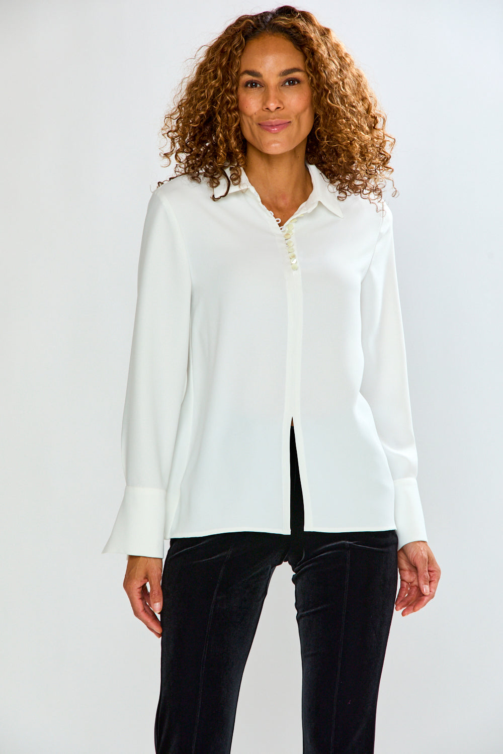 A woman with curly brown hair wearing a white long-sleeve button-up blouse with a split neckline and black pants, standing against a light gray studio background.