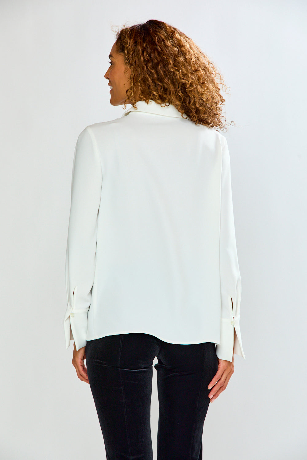 A woman with curly brown hair is shown from the back wearing a white long-sleeve blouse with side tie details and black pants, set against a plain light gray studio background.