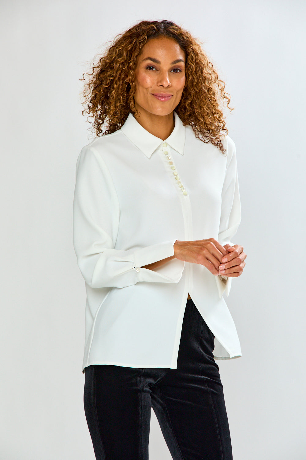 A woman with curly brown hair wearing a white long-sleeve button-up blouse with pearl-like buttons and black pants, standing against a light gray studio background.