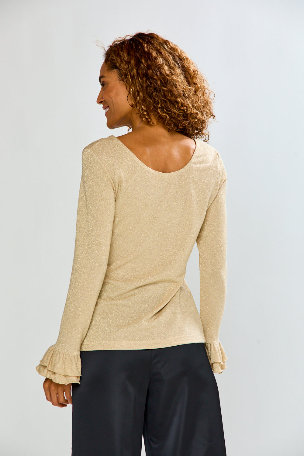 The Jonie Top - Gold in a soft knit with long sleeves and ruffled cuffs shown from the back with black pants for a chic sophisticated look