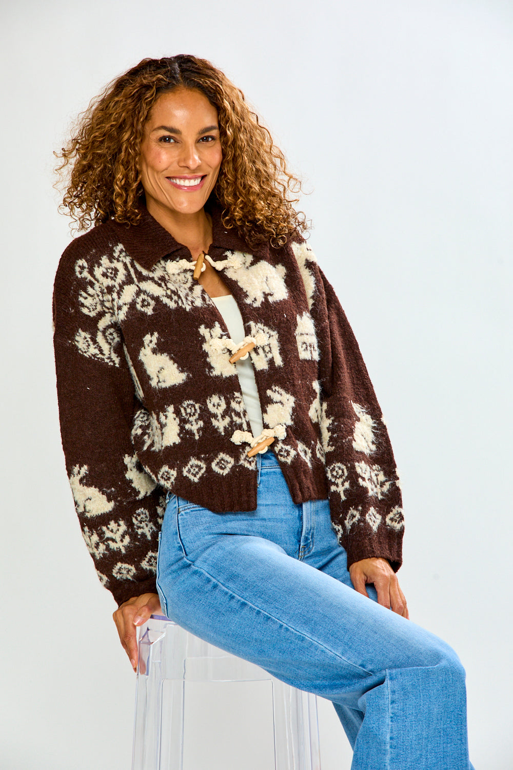 The Bunny Toggle Sweater - Brown Multi modeled by a smiling woman with curly hair and blue jeans featuring brown and cream bunny motifs and toggle closures