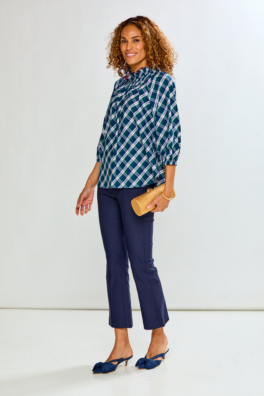 A woman modeling a blue and white plaid button-down shirt, navy blue cropped pants, and navy blue slingback heels, holding a tan clutch against a plain white studio background.