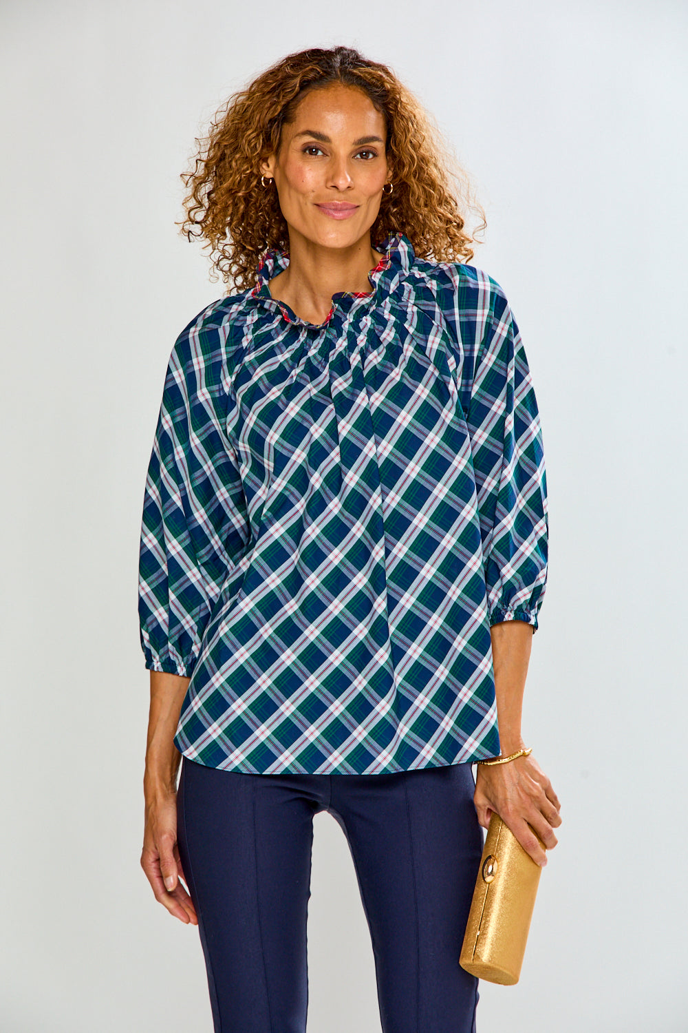 A woman with curly brown hair wearing a blue, green, and white plaid button-up shirt and navy pants, holding a gold clutch against a light gray studio background.