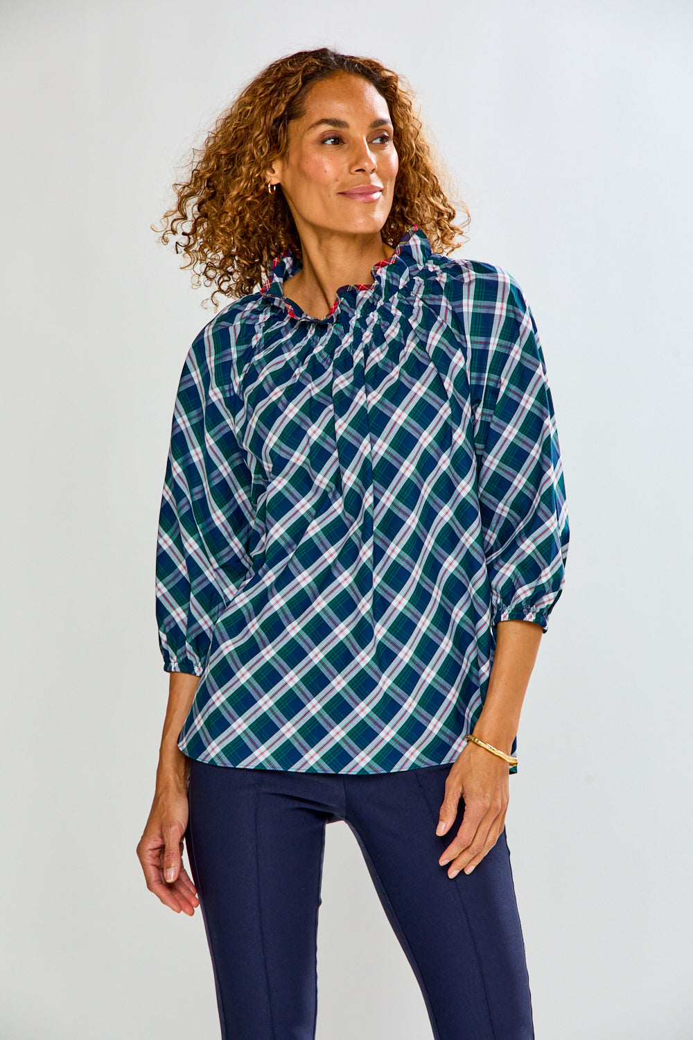 A woman modeling a long-sleeve plaid blouse with a smocked chest, in navy blue, green, and white, paired with navy pants, against a light gray studio background.