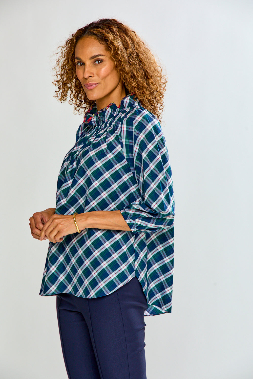 A woman with curly brown hair wearing a teal and white plaid long-sleeve blouse with dark navy pants, standing against a light gray studio background.