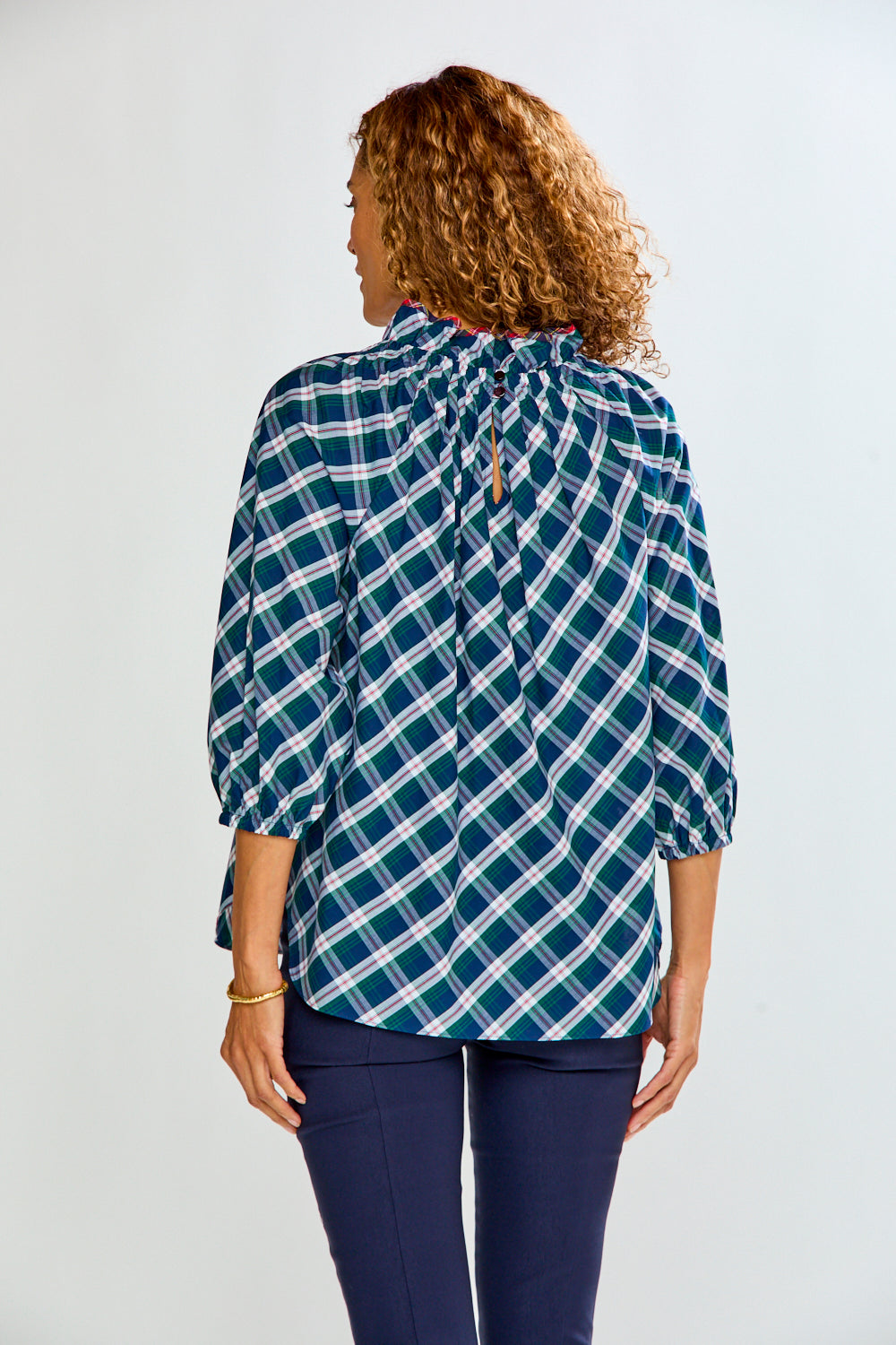 A woman viewed from the back wearing a long-sleeve plaid blouse in teal, white, and light blue, paired with navy pants, against a light gray studio background.