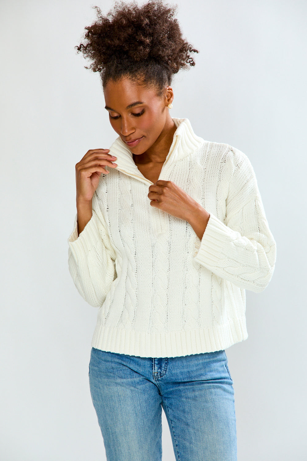 Woman wearing a white cable knit sweater and blue jeans against a plain background