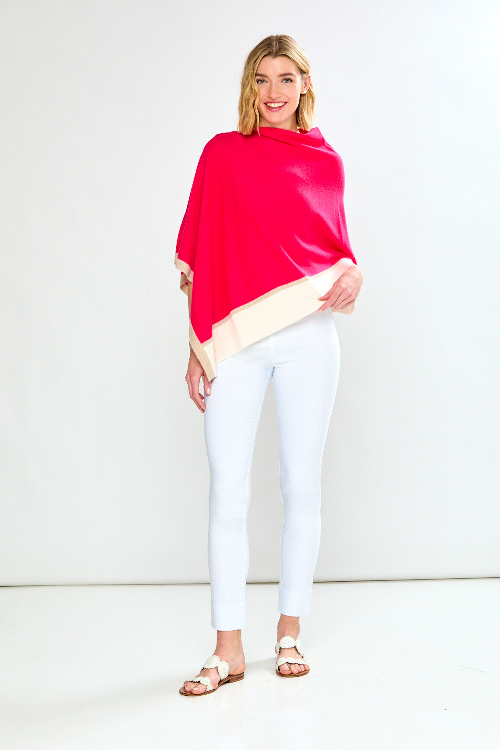 Woman wearing a bright pink poncho over a beige top with white pants on a plain background