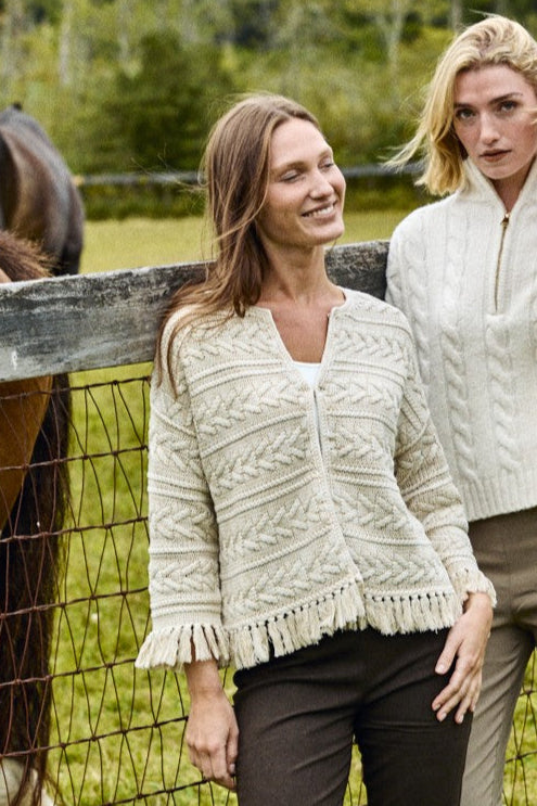Two women wearing cream cable knit cardigans standing next to a horse in a field.