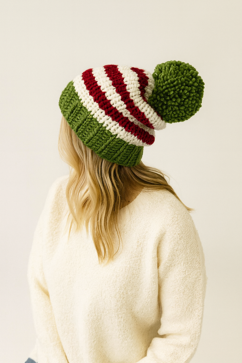 Person wearing a green and red striped knit beanie with a pom-pom on a plain background