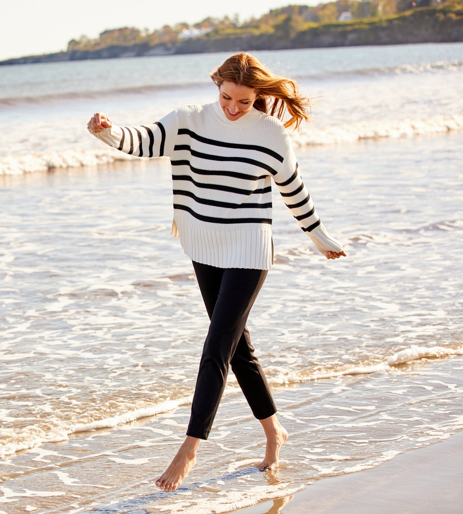 Woman walking on the beach wearing black fitted pants and an ivory and black stripe turtleneck. 