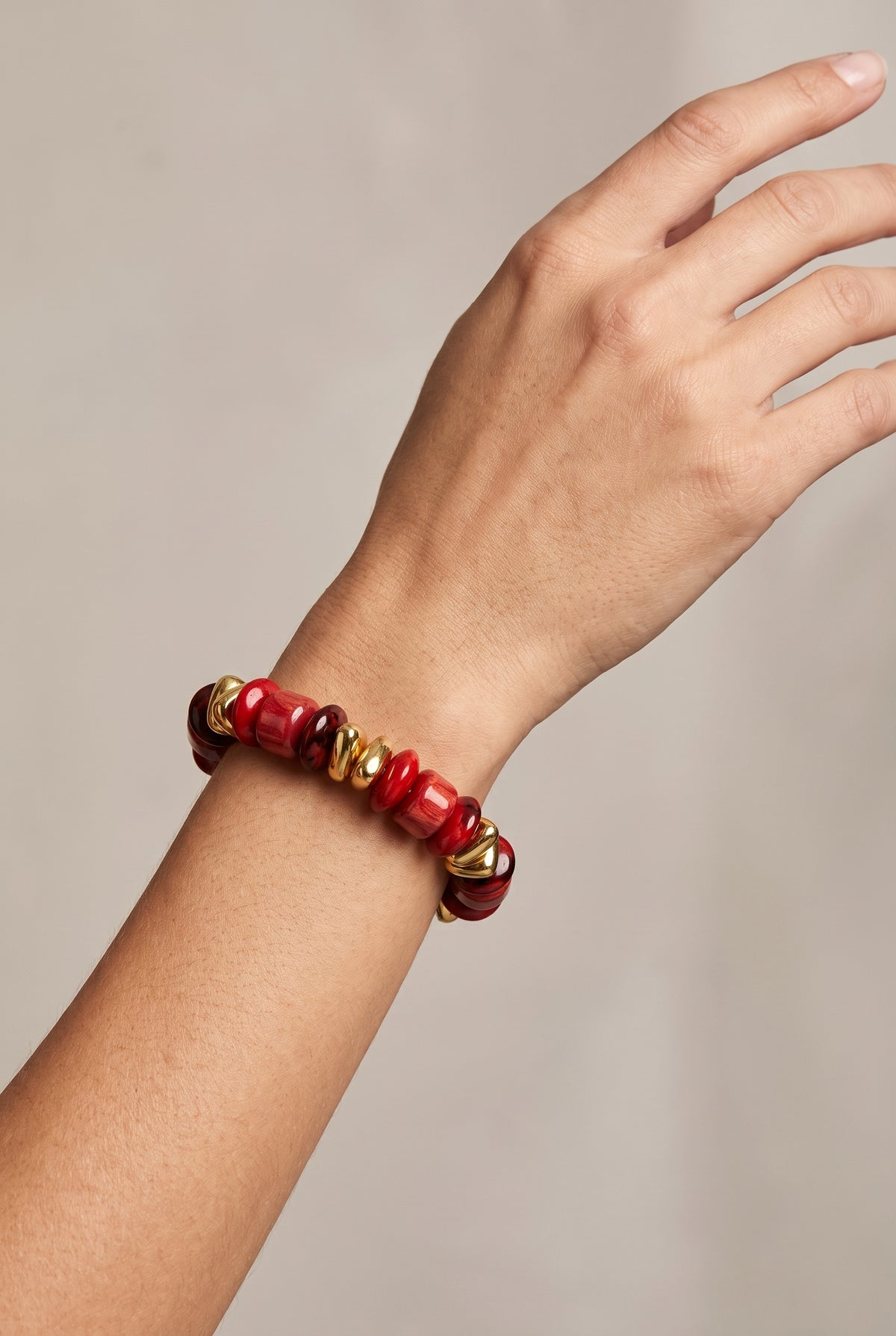 A close-up of a person's wrist wearing a beaded stretch bracelet with red and gold beads, set against a neutral light gray background.