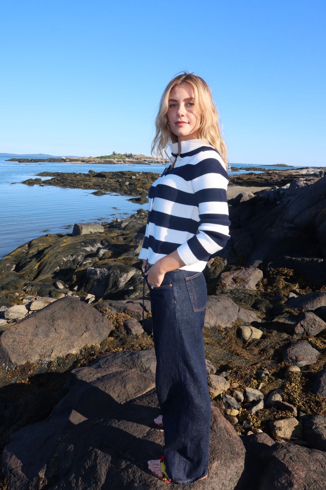 Woman in a striped sweater and jeans standing on rocks by the water