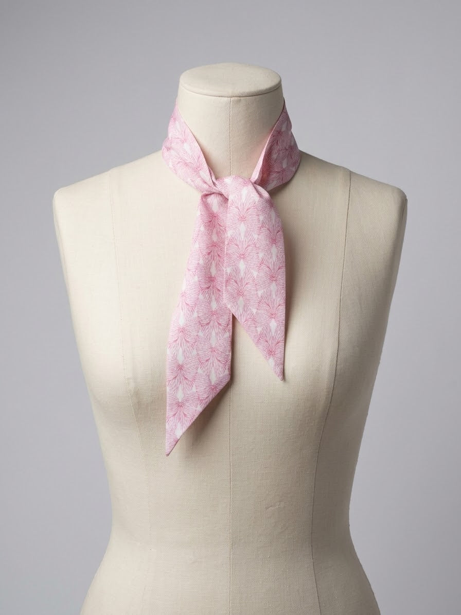 A light pink silk scarf with a white floral pattern, tied around the neck of a cream-colored fabric dress form against a neutral gray background.