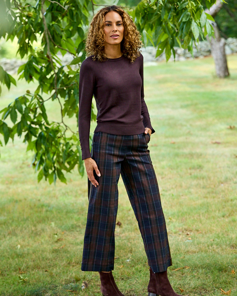 A woman stands outdoors in a grassy park area under leafy trees, wearing a dark plum long-sleeve knit top and dark plaid cropped trousers with brown ankle boots.