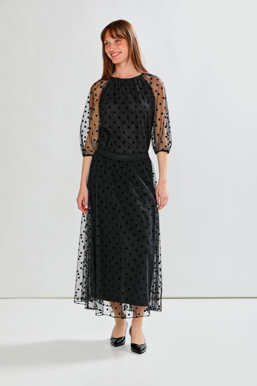 Woman wearing a black polka dot sheer top and black pants on a white background
