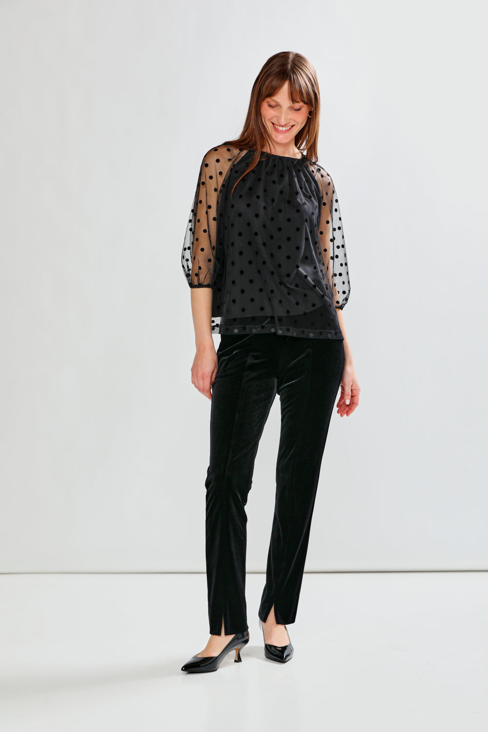Woman wearing a black polka dot sheer top and black pants on a white background