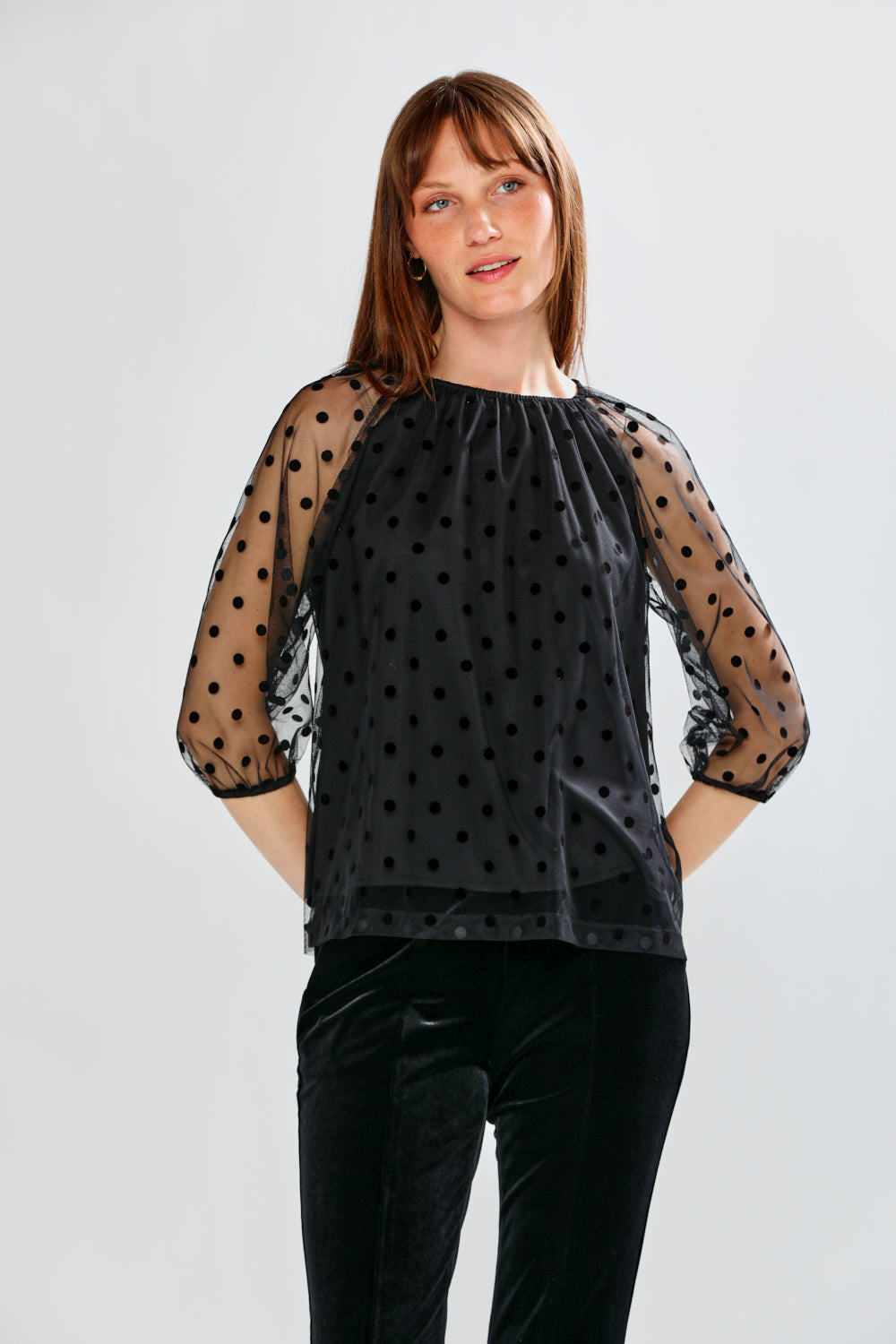 Woman wearing a black polka dot sheer top and black pants on a white background