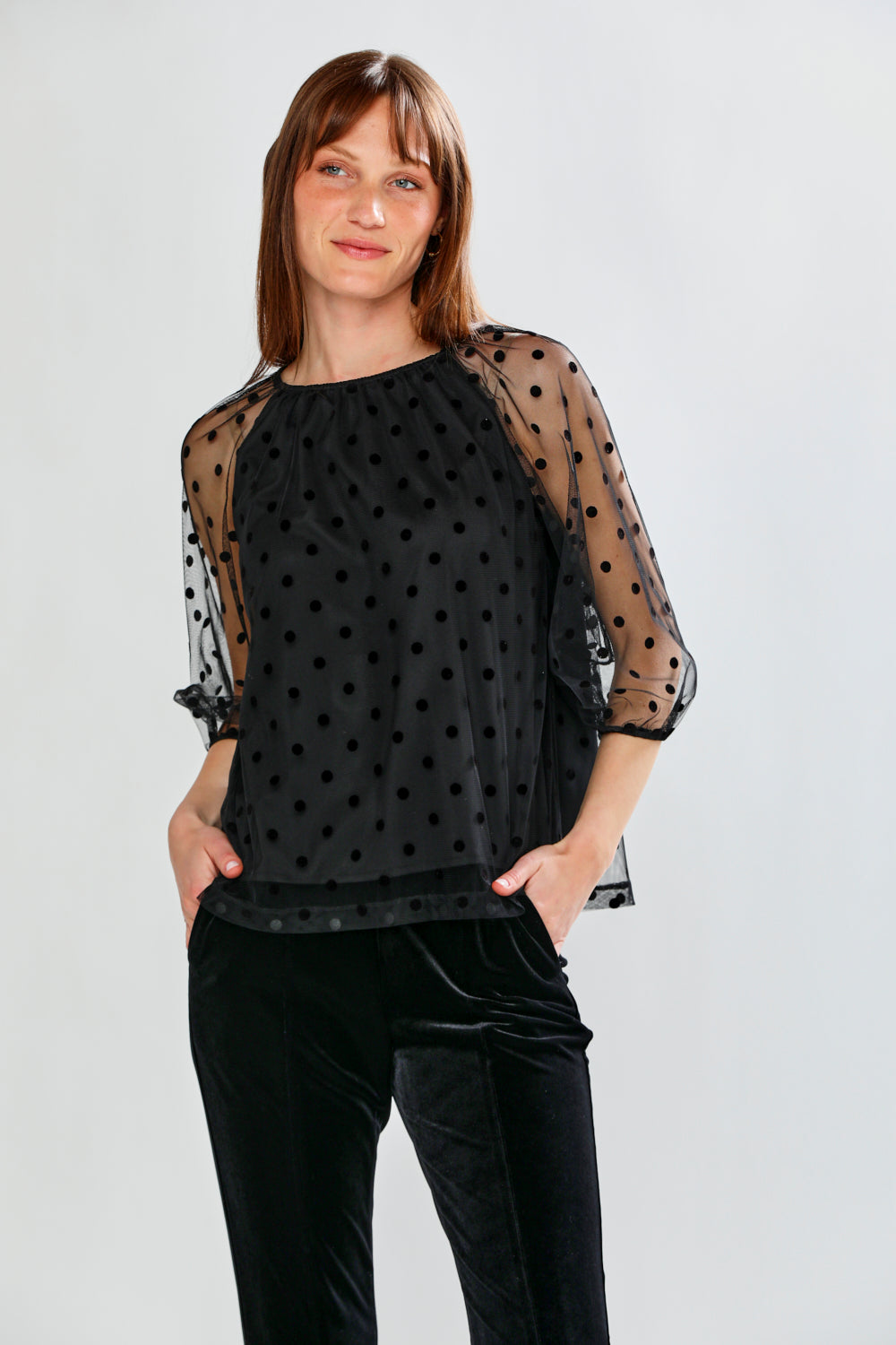 Woman wearing a black polka dot sheer top and black pants on a white background