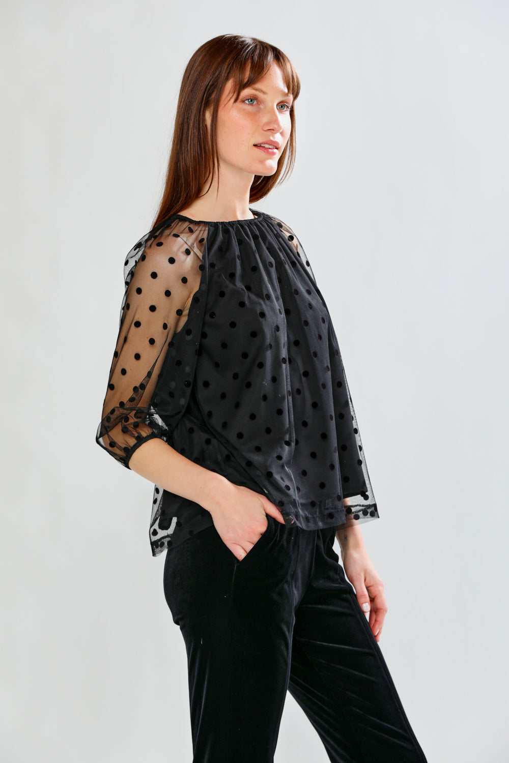 Woman wearing a black polka dot sheer top and black pants on a white background