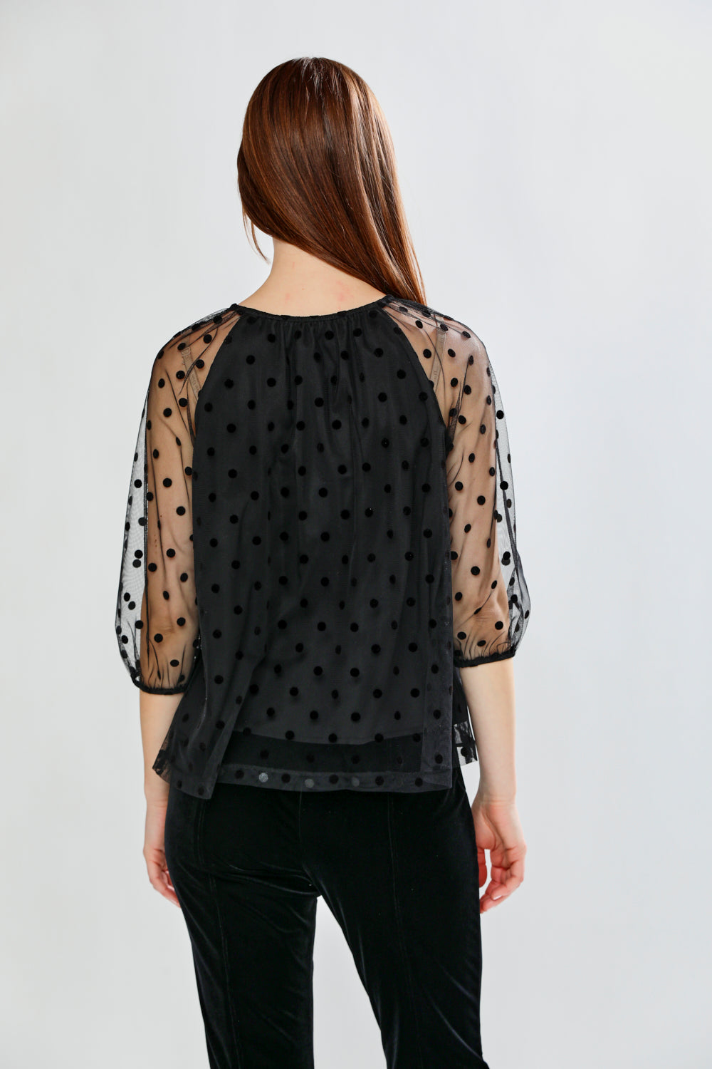 Woman wearing a black polka dot sheer top and black pants on a white background