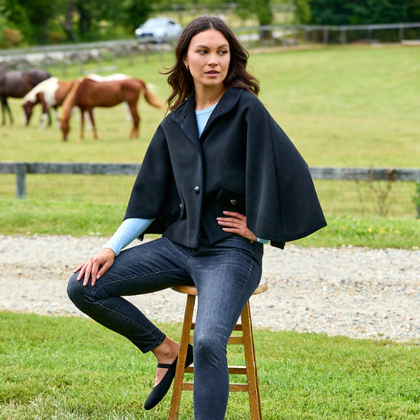 A woman sits on a wooden stool in a grassy field with horses in the background, wearing a black cape-style jacket over a light blue top and dark gray jeans.