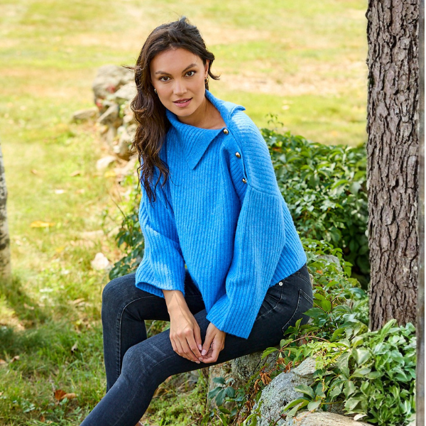 A woman sitting outdoors on a stone wall wearing a bright blue ribbed knit sweater with a high draped collar and dark jeans, with a grassy yard and tree trunk in the background.