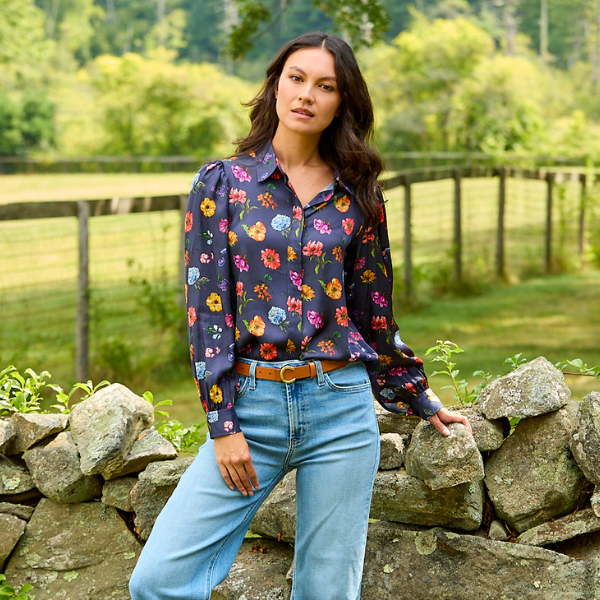 A woman stands outdoors in front of a stone wall and green field, wearing a dark navy floral long-sleeve button-up blouse with light blue jeans and a brown belt.