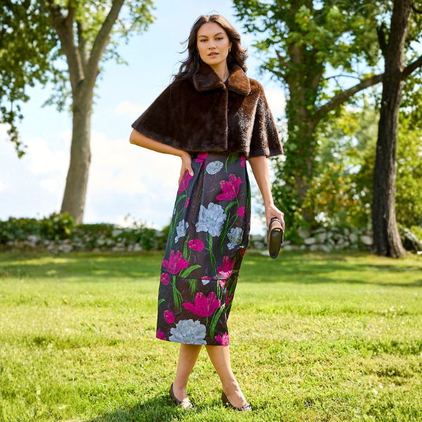 A woman stands on a grassy lawn outdoors wearing a dark brown faux-fur cropped cape jacket and a dark midi skirt with a pink, purple, and white floral print. She is holding a brown leather clutch.